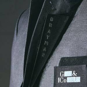 Angled close-up frontal shot of Grayman and Company branded velcro lapels sitting on a grey herringbone suit jacket