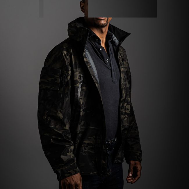 redacted man in studio wearing light grey merino pique polo shirt by Grayman and Company and multicam black jacket by OTTE Gear with grey backdrop
