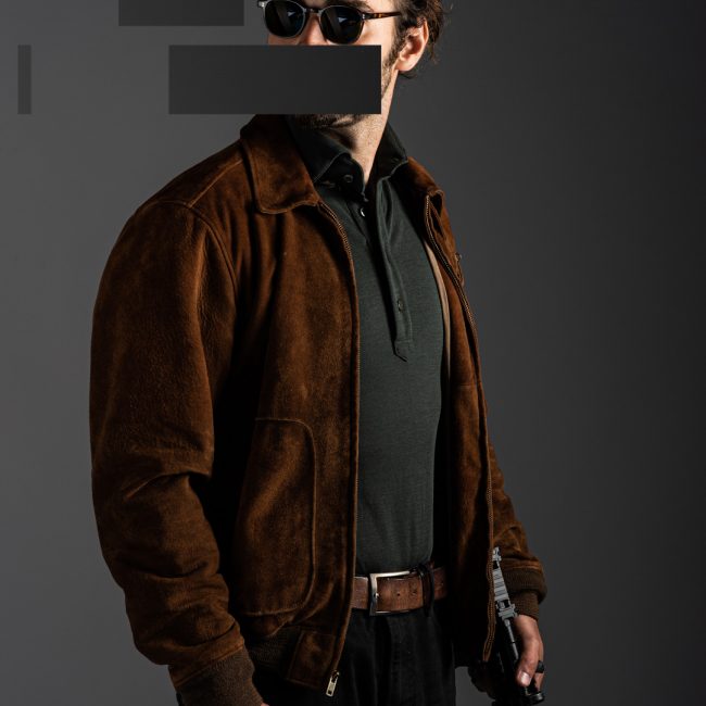redacted man in studio wearing green merino pique polo shirt, ballistic wayfarer sunglasses and suede jacket by Grayman and Company with grey backdrop