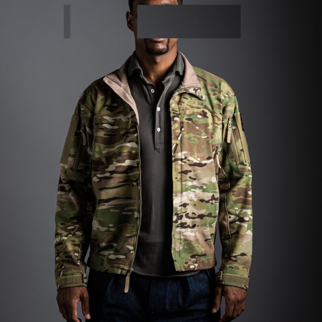 model wearing Grayman and Company pique merino polo shirt in taupe colour with massif multicam jacket against grey background with redacted face