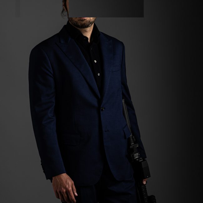 redacted man with rifle and Cloud Defensive light in studio wearing navy blue merino pique polo shirt and blue tactical suit by Grayman and Company with grey backdrop