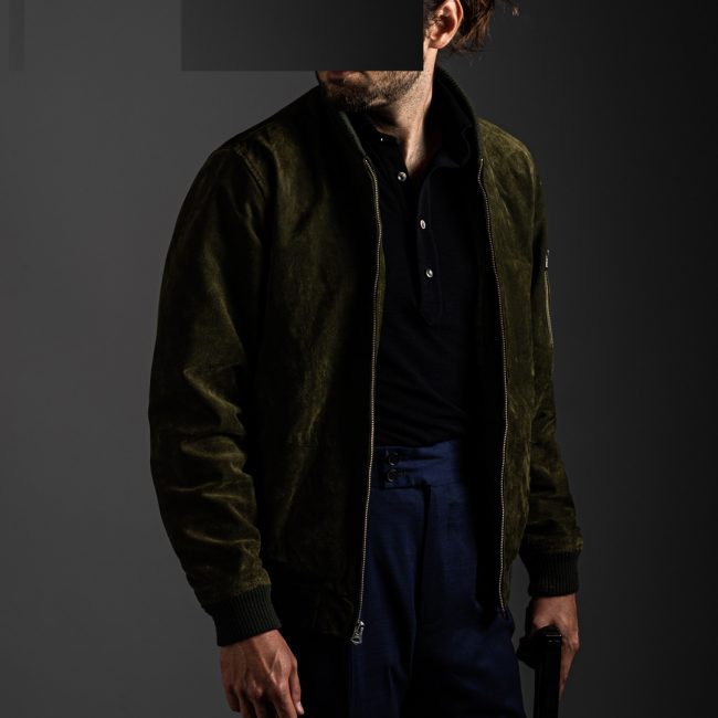 redacted man with GLock pistol in studio wearing navy blue merino pique polo shirt by Grayman and Company and green suede jacket with grey backdrop