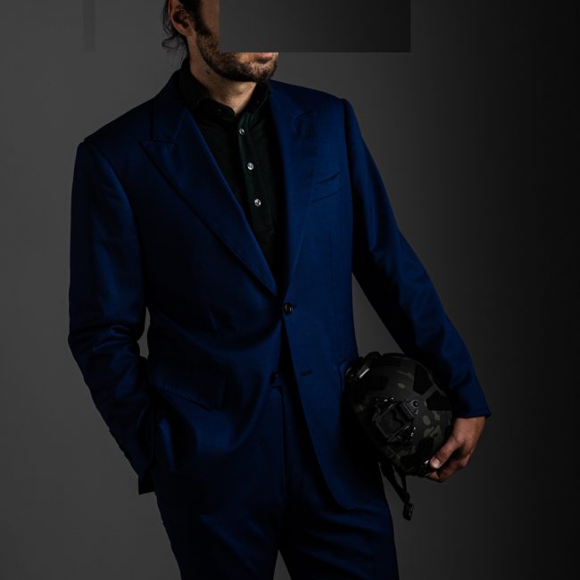 redacted man in studio wearing dark grey merino pique polo shirt and blue tactical suit by Grayman and Company with grey backdrop