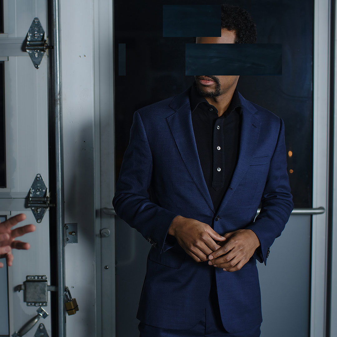 Man with redacted face wearing Grayman and Company pique merino polo shirt and blue suit in front of warehouse door